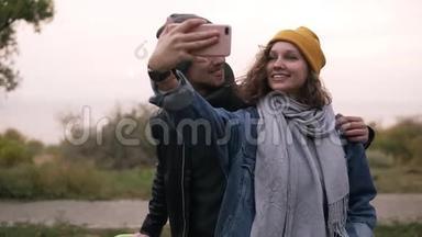 快乐的年轻夫妇在秋天公园用手机自拍。 戴着黄色帽子的女孩拿着智能手机。 美丽美丽
